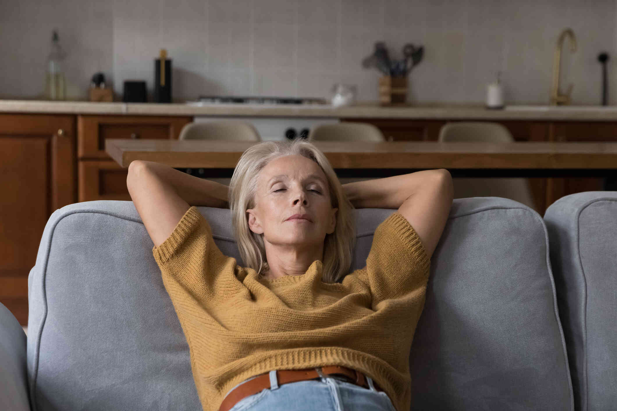 A middle aged woman in a yellow sweater reclines on the couch with her eyes closed and her hands behind her head.